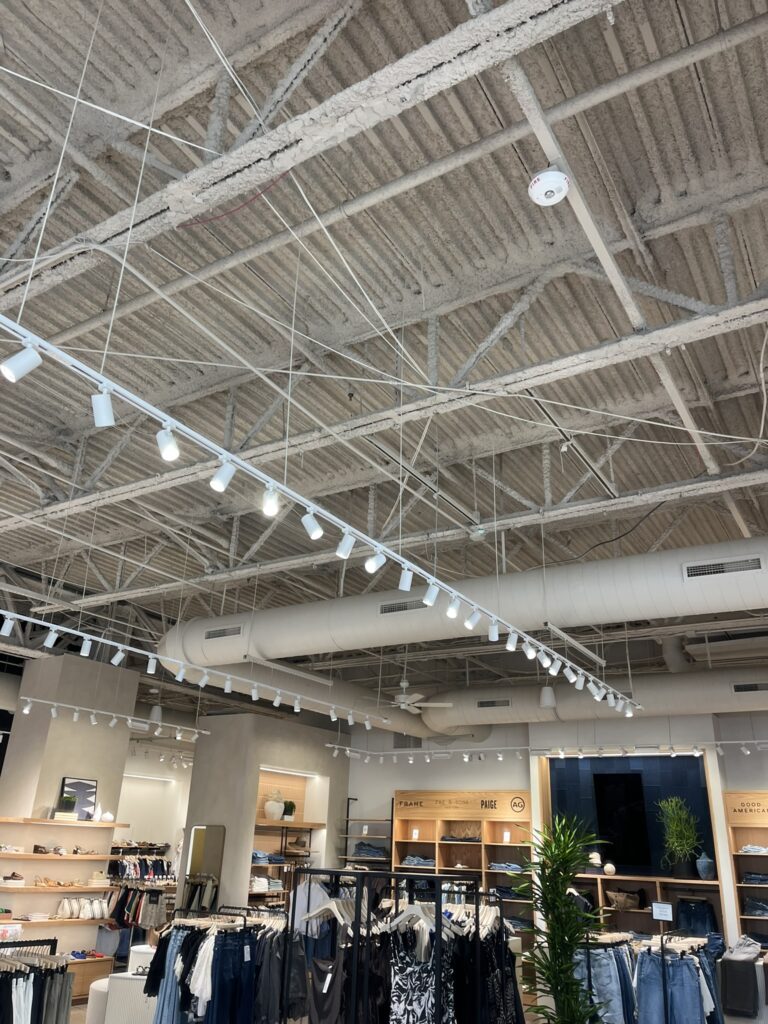 Large white round ductwork runs across an exposed ceiling in a commercial retail store, with track lighting and clothing racks below.