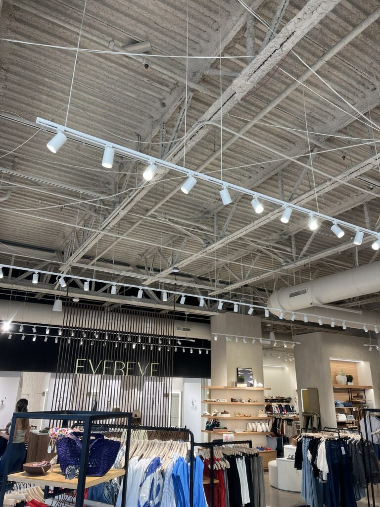 Exposed ceiling in a retail store with a large, insulated round HVAC supply duct, track lighting, and clothing displays.
