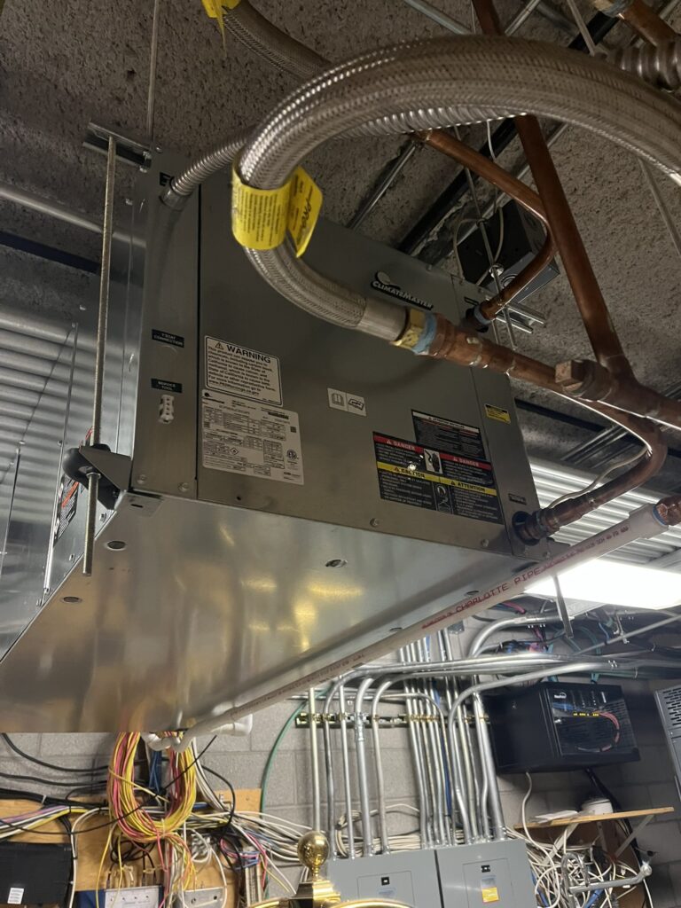 A new ClimateMaster water source heat pump unit suspended from a ceiling, with various pipes and electrical conduits connected.