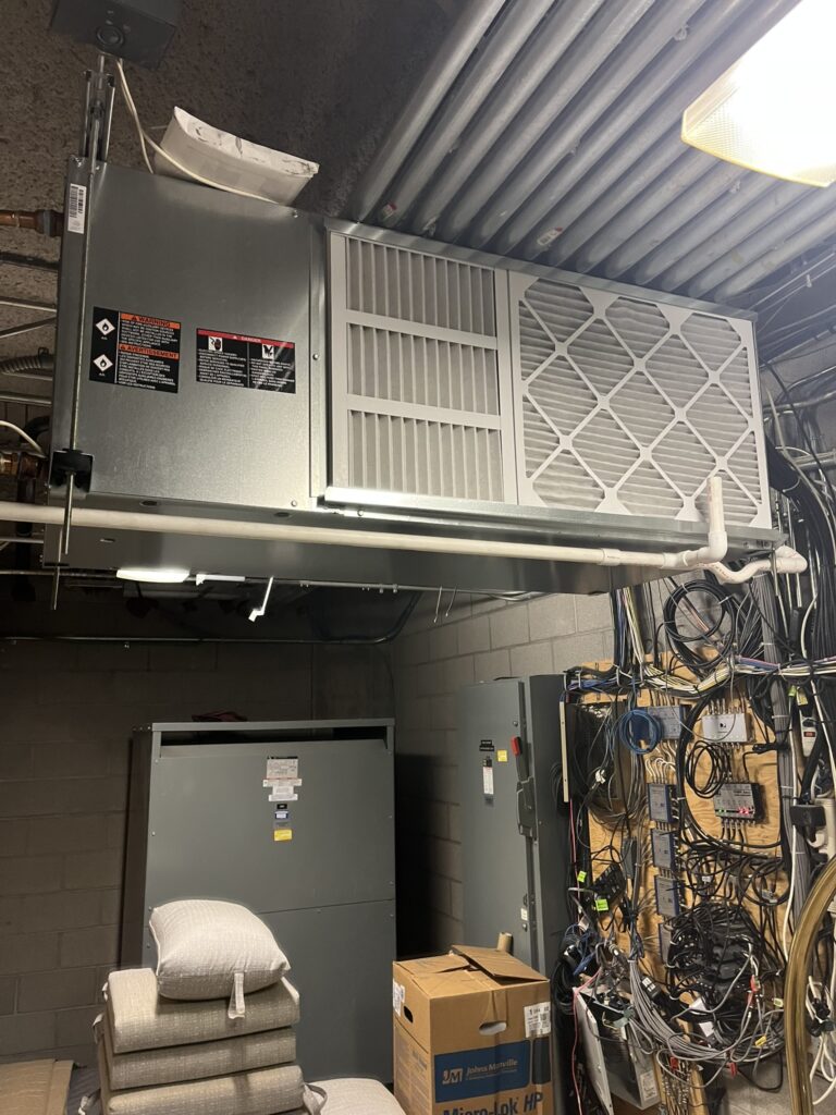 New water source heat pump air handler suspended from a ceiling in a commercial mechanical room, with other HVAC units below.
