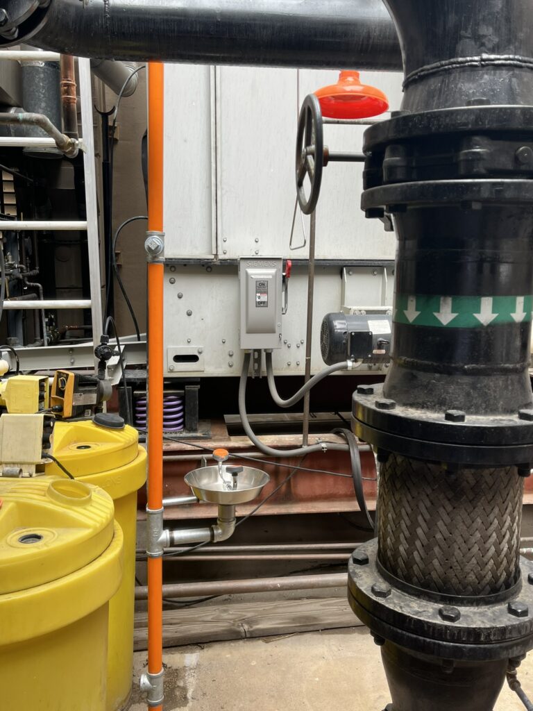 An eyewash station and large industrial pipes in a commercial mechanical room, with a large HVAC unit in the background.