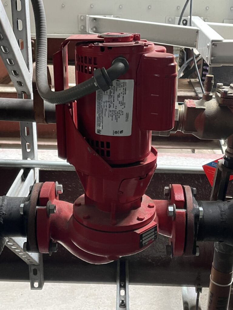 A large red commercial booster pump is installed on black pipes with flanges, supported by metal racking in a mechanical room.