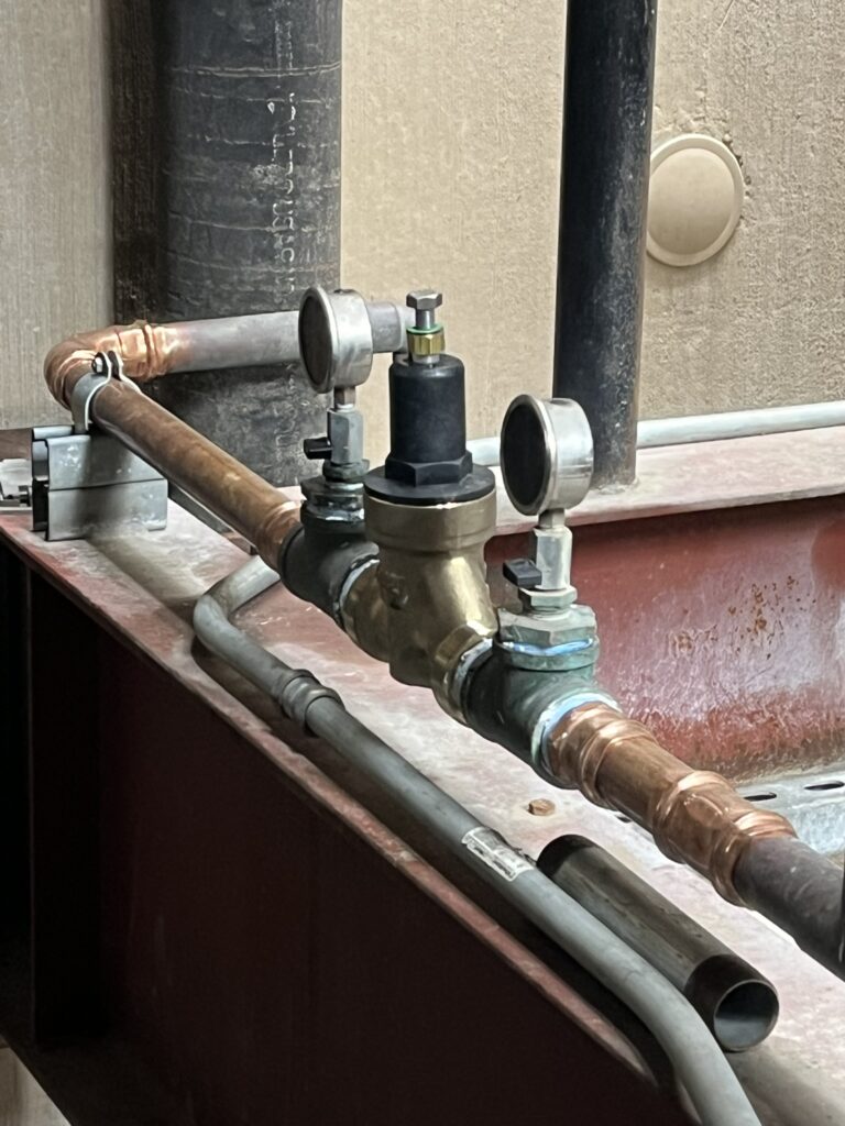 Close-up of copper plumbing with a brass valve assembly and two pressure gauges on a red metal beam.