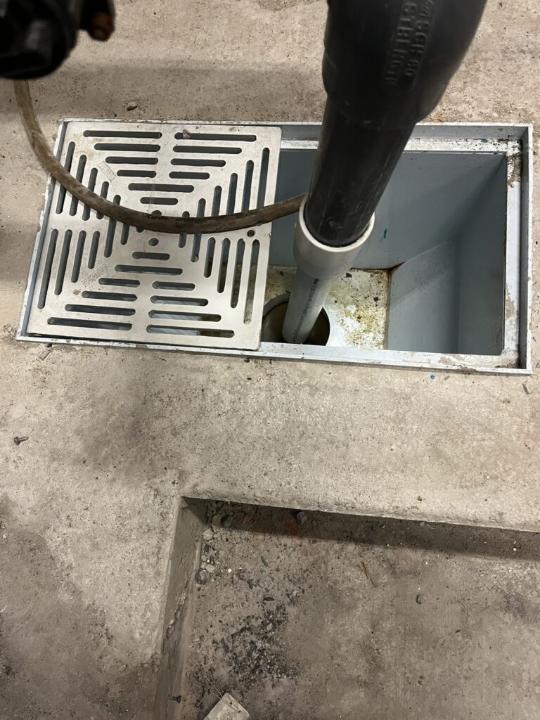 A floor drain with a metal grate and two pipes inserted into it, set in a concrete floor in a commercial mechanical room.