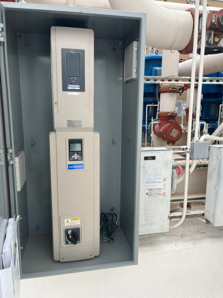 Two Yaskawa variable frequency drives installed in an open electrical enclosure, with commercial HVAC pipes and pumps in the background.