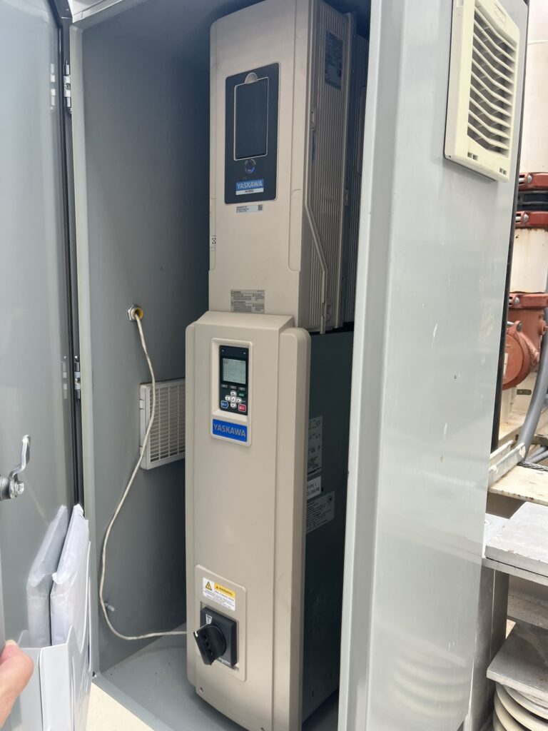 Open electrical cabinet showing two stacked Yaskawa variable frequency drives with a digital display and rotary switch.