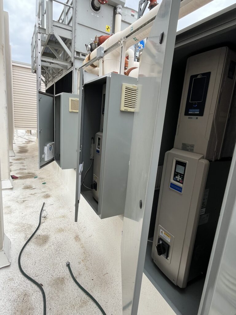 Open electrical cabinets on a commercial rooftop showing installed Yaskawa variable frequency drives next to a large commercial HVAC unit.