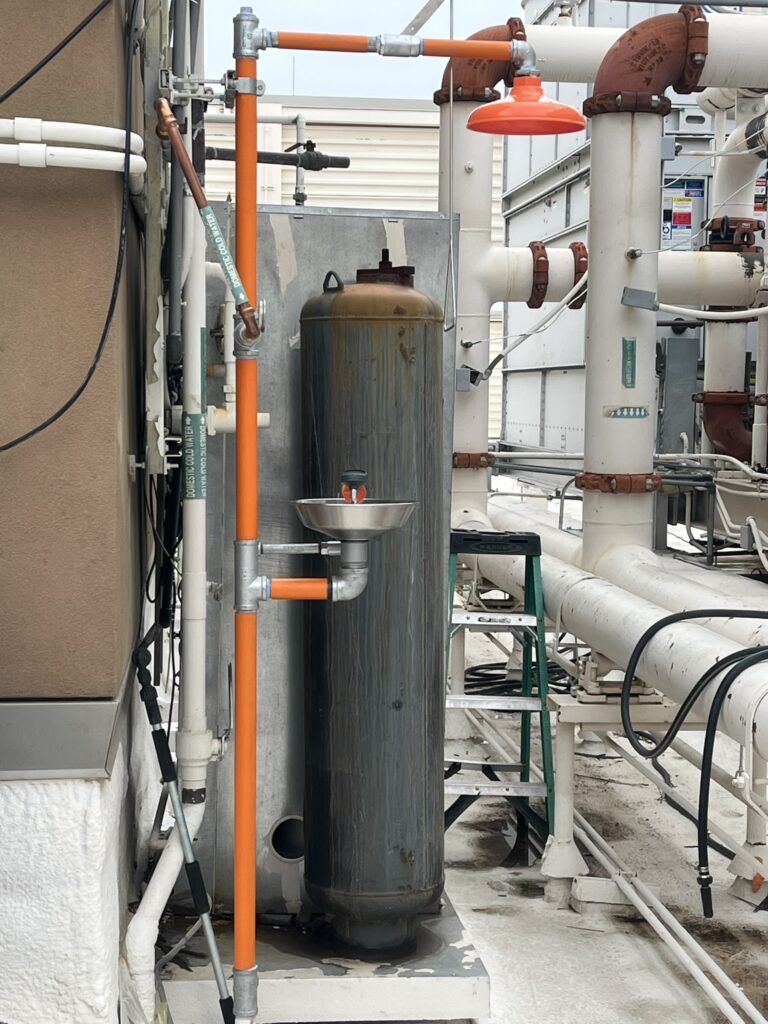An emergency eyewash and shower station with orange pipes on a commercial rooftop, surrounded by large white pipes and a rusty tank.