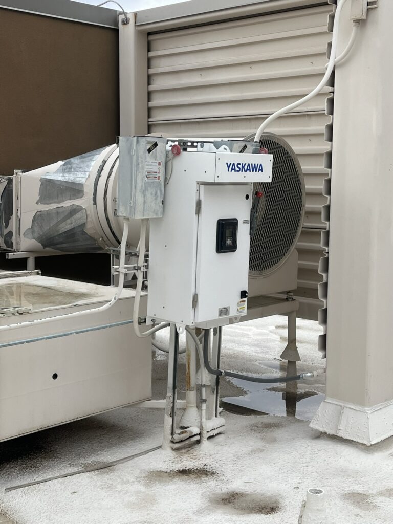 A Yaskawa variable frequency drive (VFD) control panel and a large supply fan unit with ductwork on a commercial rooftop.