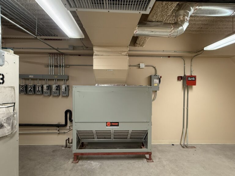 A newly installed gray Trane air handler unit in a commercial mechanical room, connected to ductwork and electrical systems.