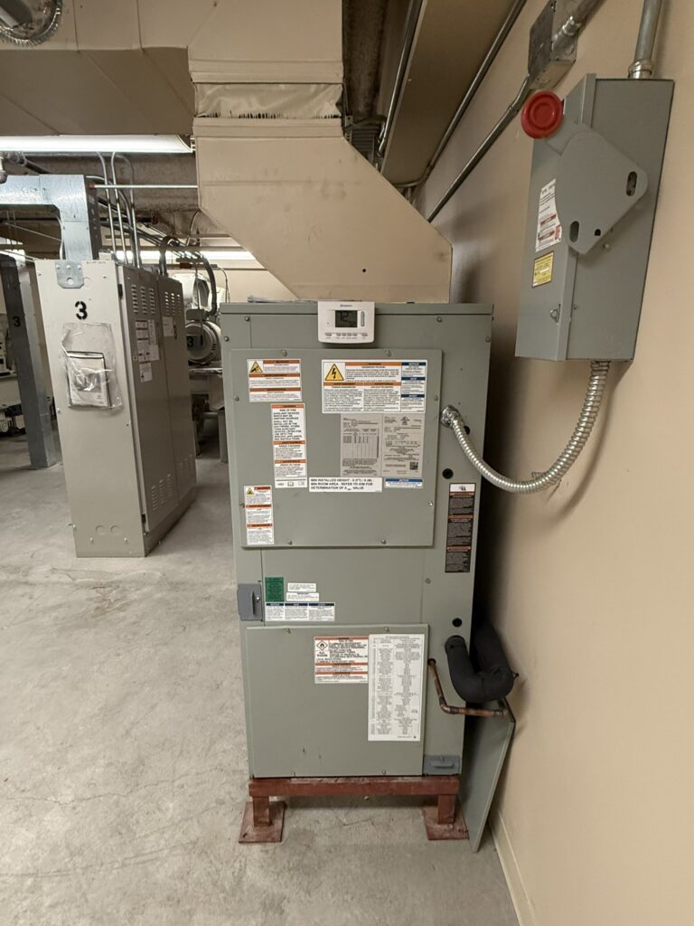 A large gray Trane air handler unit stands on a metal frame in a mechanical room, connected to overhead ductwork.