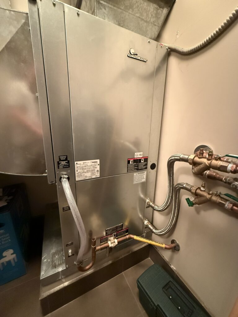 A newly installed Climatemaster water source heat pump unit with ductwork, copper pipes, and a drain line in an indoor utility space.