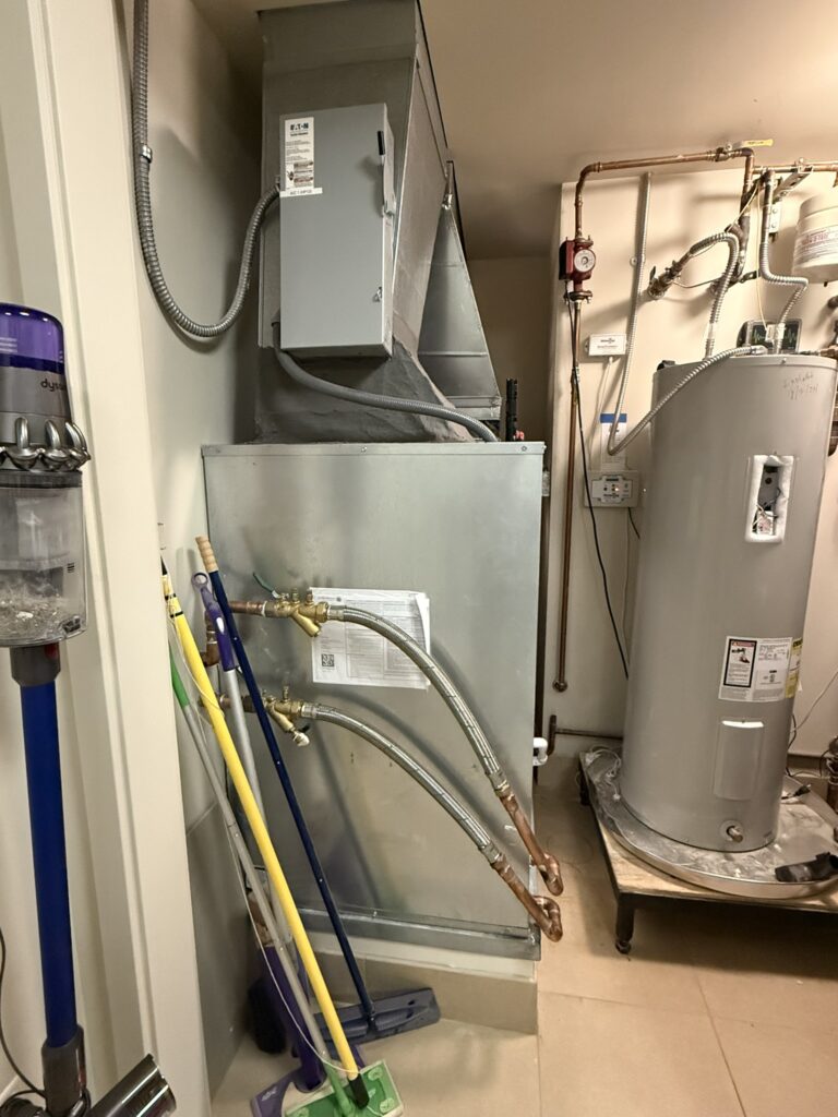 A newly installed water source heat pump unit with ductwork and flexible water lines in a utility closet, next to a water heater.