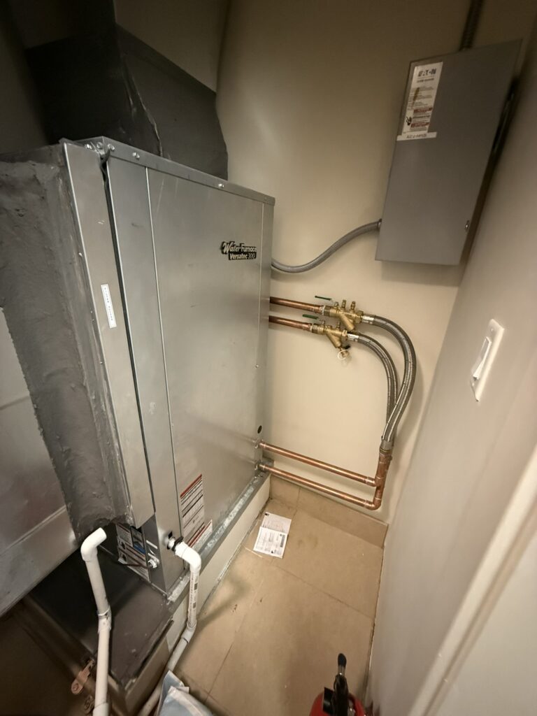 A newly installed Water Furnace Versatec 300 water-source heat pump unit in a utility closet, connected to ductwork and copper pipes.