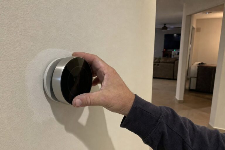 A hand adjusts a modern smart thermostat mounted on a light-colored wall in a home, with a blurred living area in the backgro