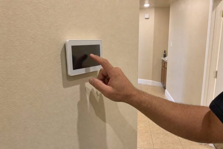 A hand touches the screen of a white smart thermostat on a beige wall in a home hallway.