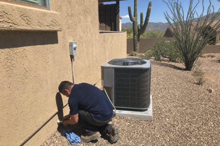HVAC technician kneeling to work on a condensate drain line next to an outdoor AC condenser unit at a desert home.