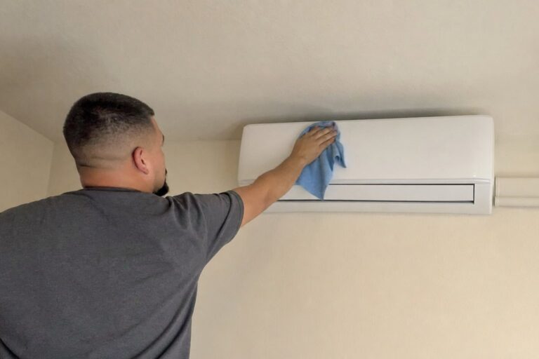 A technician in a grey shirt wipes down a white wall-mounted mini-split air conditioning unit with a blue cloth in an indoor setting.