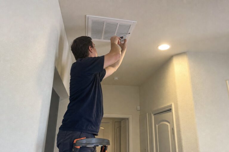 A technician on a ladder uses a screwdriver to work on a white ceiling air return vent in a residential home.
